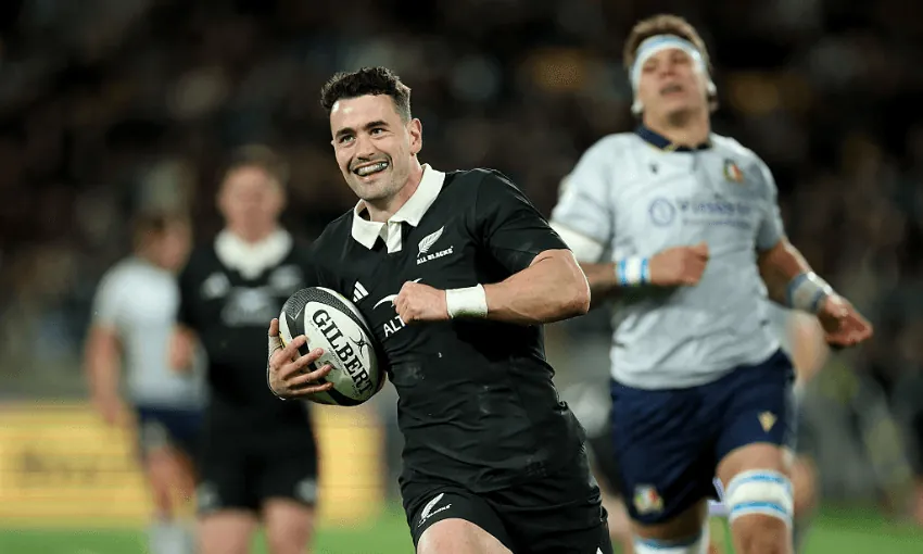 a rugby player in an all blacks uniform smiles while running with the ball as a player in a grey uniform chases behind